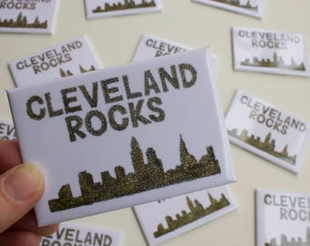 May include: A collection of white square magnets with the text "Cleveland Rocks" and a black and white silhouette of the Cleveland skyline. The skyline is made up of small, textured circles.