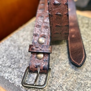 May include: A brown leather belt featuring a textured crocodile-skin pattern, a rectangular metal buckle, and two circular metal rivets securing the strap near the buckle. The belt has a pointed tip and visible stitching along the edges.