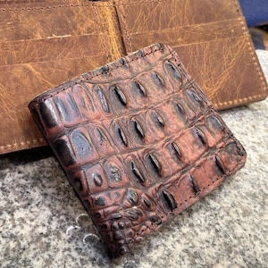 May include: A bi-fold wallet featuring a textured, dark brown crocodile-embossed leather exterior and a smooth, light brown distressed leather interior with visible stitching along the edges and multiple internal card slots.
