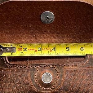 May include: A brown leather pouch with a woven pattern and two snap closures. The pouch is measured with a tape measure, showing it is 7 inches long.