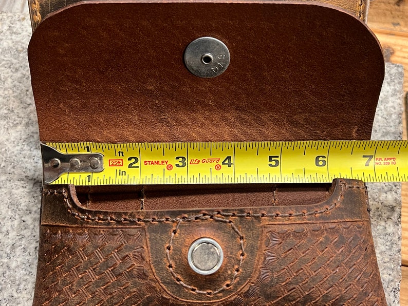 May include: A brown leather pouch with a woven pattern and two snap closures. The pouch is measured with a tape measure, showing it is 7 inches long.