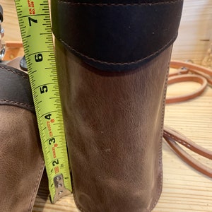 May include: A brown leather cylindrical case with a dark brown leather flap and a white leather trim. The case is being measured with a yellow tape measure that reads 8 inches.