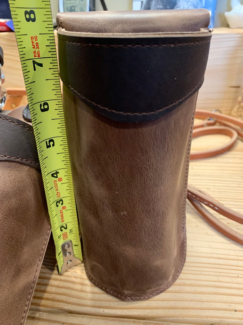May include: A brown leather cylindrical case with a dark brown leather flap and a white leather trim. The case is being measured with a yellow tape measure that reads 8 inches.
