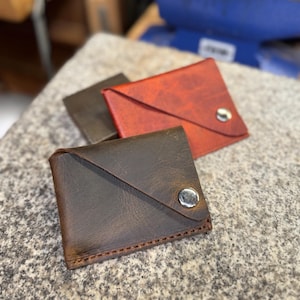 May include: Three leather wallets in shades of brown and red are displayed on a textured surface. The wallets have a minimalist design with a flap closure secured by a silver snap. The wallets are likely handmade.