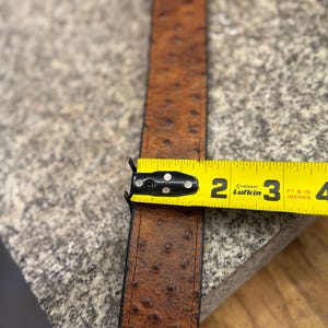 May include: A brown belt with a textured ostrich-style pattern and stitched edges. A yellow Lufkin measuring tape is positioned across the belt, showing a width of approximately 1.5 inches. The belt features a dark brown finish with darker spots.