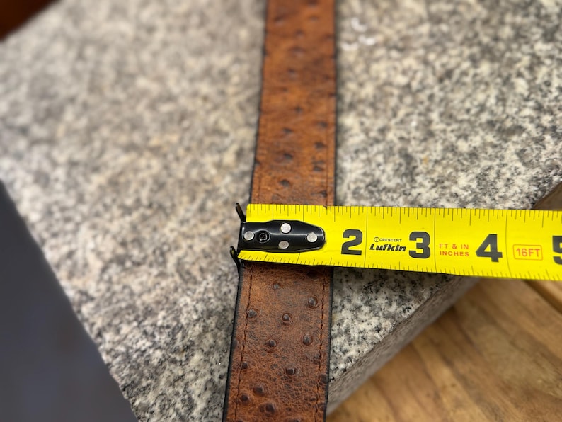 May include: A brown belt with a textured ostrich-style pattern and stitched edges. A yellow Lufkin measuring tape is positioned across the belt, showing a width of approximately 1.5 inches. The belt features a dark brown finish with darker spots.