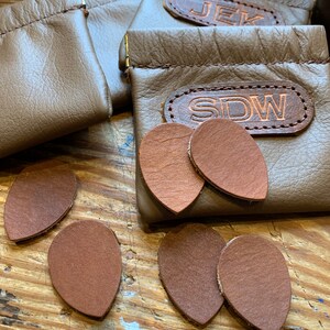 Peut inclure: Cinq m&eacute;diators en cuir marron, deux sur une surface en bois et trois sur une pochette en cuir marron avec les initiales "SDW" brod&eacute;es dessus. Deux autres pochettes sont visibles en arri&egrave;re-plan, l'une avec les initiales "JEK" brod&eacute;es dessus.