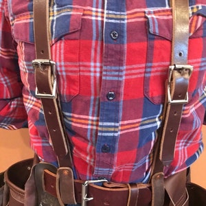 May include: A brown leather tool belt with suspenders. The belt has a silver buckle and multiple pockets. The suspenders are attached to the belt and have silver buckles. The person wearing the belt is wearing a red and blue checked shirt and blue jeans.