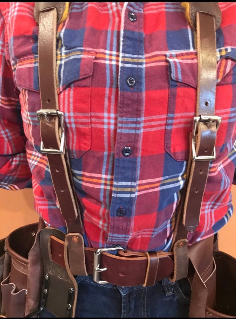May include: A brown leather tool belt with suspenders. The belt has a silver buckle and multiple pockets. The suspenders are attached to the belt and have silver buckles. The person wearing the belt is wearing a red and blue checked shirt and blue jeans.