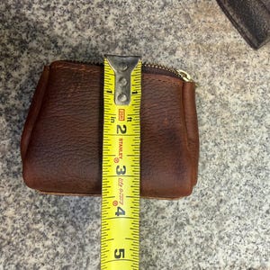 May include: A brown leather coin purse with a gold zipper, measuring approximately 4 inches in height. The purse is made of textured leather and is shown with a yellow measuring tape.