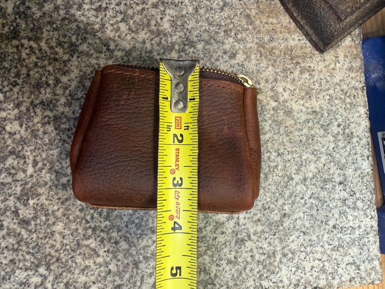May include: A brown leather coin purse with a gold zipper, measuring approximately 4 inches in height. The purse is made of textured leather and is shown with a yellow measuring tape.