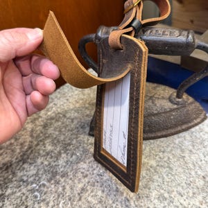 Op de afbeelding: Bruin leren bagagelabel met een rechthoekige vorm en een gestikte rand. Het label heeft een blanco witte ruimte voor naam, adres en telefoonnummer. Een leren riem en een messing gesp zijn bevestigd.