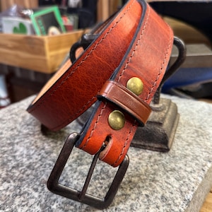 Puede incluir: Un cinturón de cuero marrón con una hebilla marrón oscuro y detalles en latón. El cinturón está doblado por la mitad y la hebilla es visible.