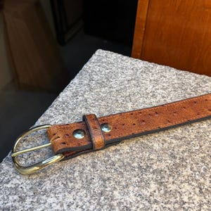 May include: A tan leather belt with a textured, dotted surface pattern. The belt features a polished gold-tone metal buckle and two matching gold-tone metal rivets securing the leather loop near the buckle. The edges are finished in dark brown.