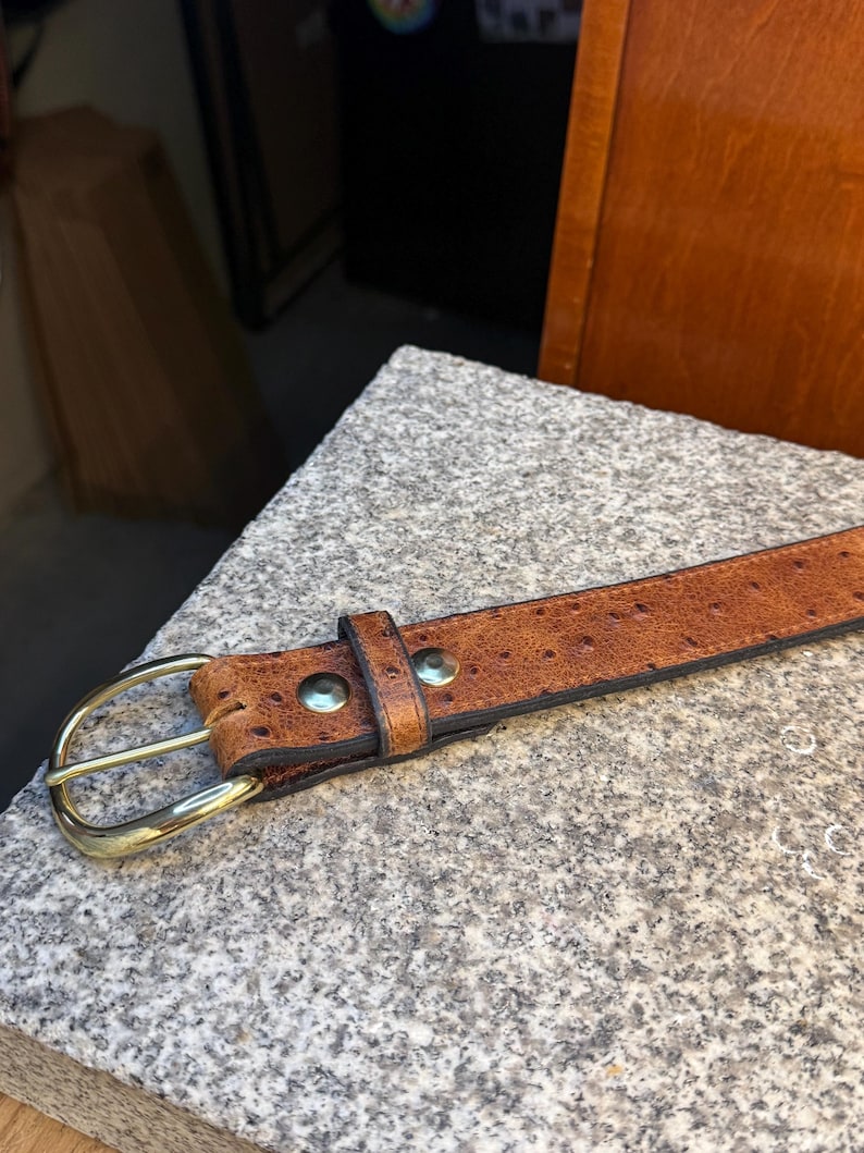 May include: A tan leather belt with a textured, dotted surface pattern. The belt features a polished gold-tone metal buckle and two matching gold-tone metal rivets securing the leather loop near the buckle. The edges are finished in dark brown.