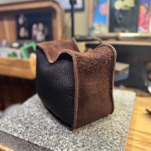 May include: A handcrafted leather pouch with a two-tone design. The bottom half is black, and the top and edges are brown. The pouch is sitting on a gray stone surface, showcasing the texture and craftsmanship of the leather.