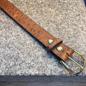 May include: A tan belt with a textured ostrich-like pattern, featuring a polished gold-tone metal buckle and two matching gold-tone metal rivets securing the leather keeper loop near the buckle end of the strap.