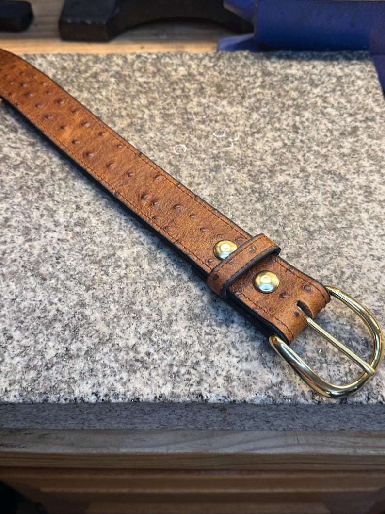 May include: A tan belt with a textured ostrich-like pattern, featuring a polished gold-tone metal buckle and two matching gold-tone metal rivets securing the leather keeper loop near the buckle end of the strap.