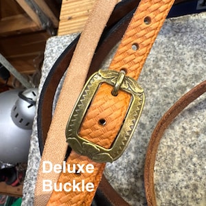 May include: Close-up of a tan leather belt with a decorative brass buckle. The belt has a woven texture and is labeled "Deluxe Buckle". Other belts in various colors are visible in the background.