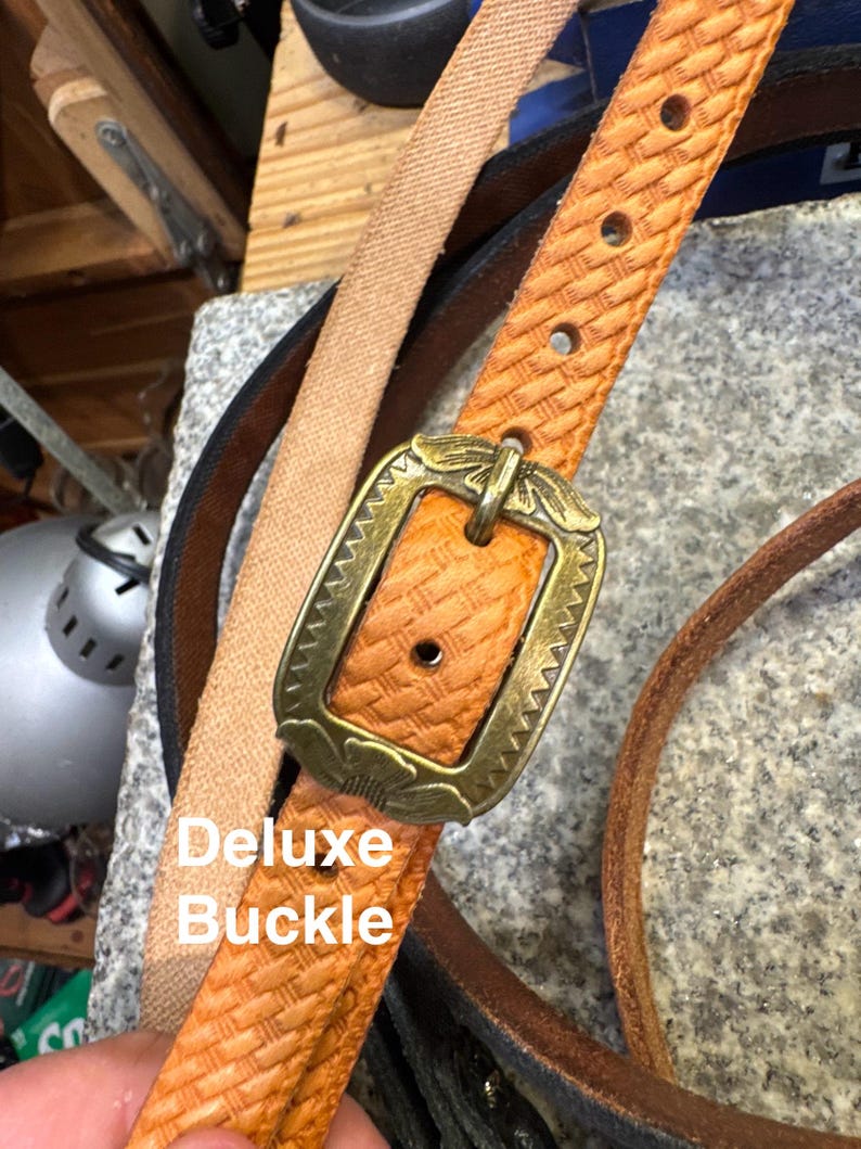 May include: Close-up of a tan leather belt with a decorative brass buckle. The belt has a woven texture and is labeled "Deluxe Buckle". Other belts in various colors are visible in the background.