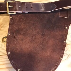 May include: A brown leather tool belt with a suede pouch and brass rivets. The belt has a brass buckle and a pointed flap.