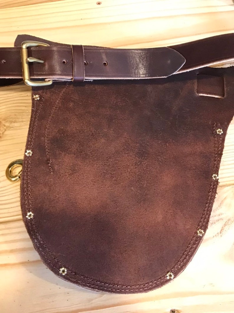 May include: A brown leather tool belt with a suede pouch and brass rivets. The belt has a brass buckle and a pointed flap.