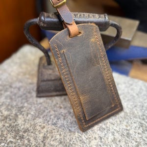 Op de afbeelding: Een bruine leren bagagelabel met een gestikte rand en een gespsluiting. Het label is rechthoekig met een kleinere rechthoekige zak aan de voorkant. De riem en de gesp zijn lichtbruin.