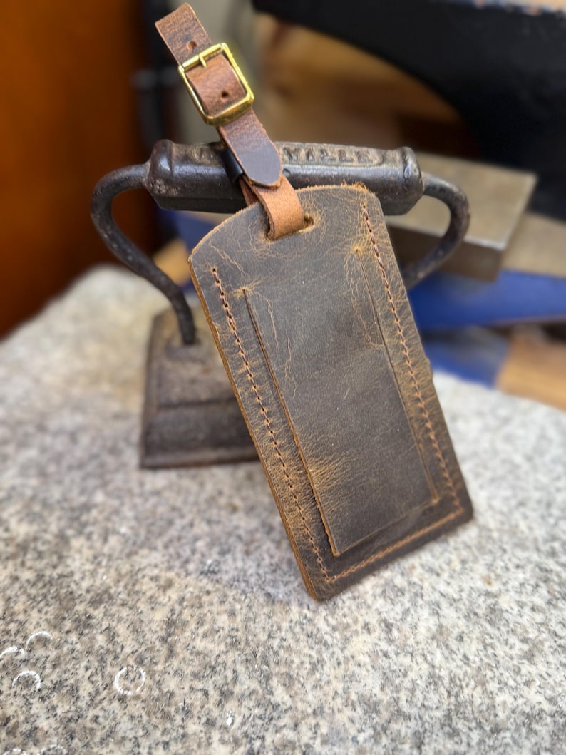 Op de afbeelding: Een bruine leren bagagelabel met een gestikte rand en een gespsluiting. Het label is rechthoekig met een kleinere rechthoekige zak aan de voorkant. De riem en de gesp zijn lichtbruin.