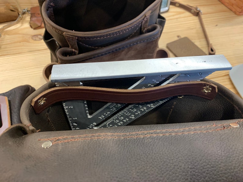May include: A brown leather tool bag with a brown leather strap and a silver metal ruler inside.