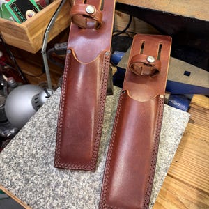 Könnte beinhalten: Zwei braune Lederscheiden mit Gürtelschlaufen und Druckknöpfen. Die Scheiden sind rechteckig mit abgerundeten Böden und genähten Kanten. Die Gürtelschlaufen sind mit silberfarbenen Druckknöpfen befestigt. Das Leder hat einen satten, rotbraunen Farbton.