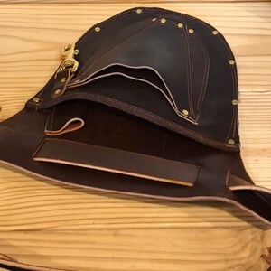 May include: A brown leather tool belt with multiple pockets and a brass buckle. The belt is open and the inside is visible.