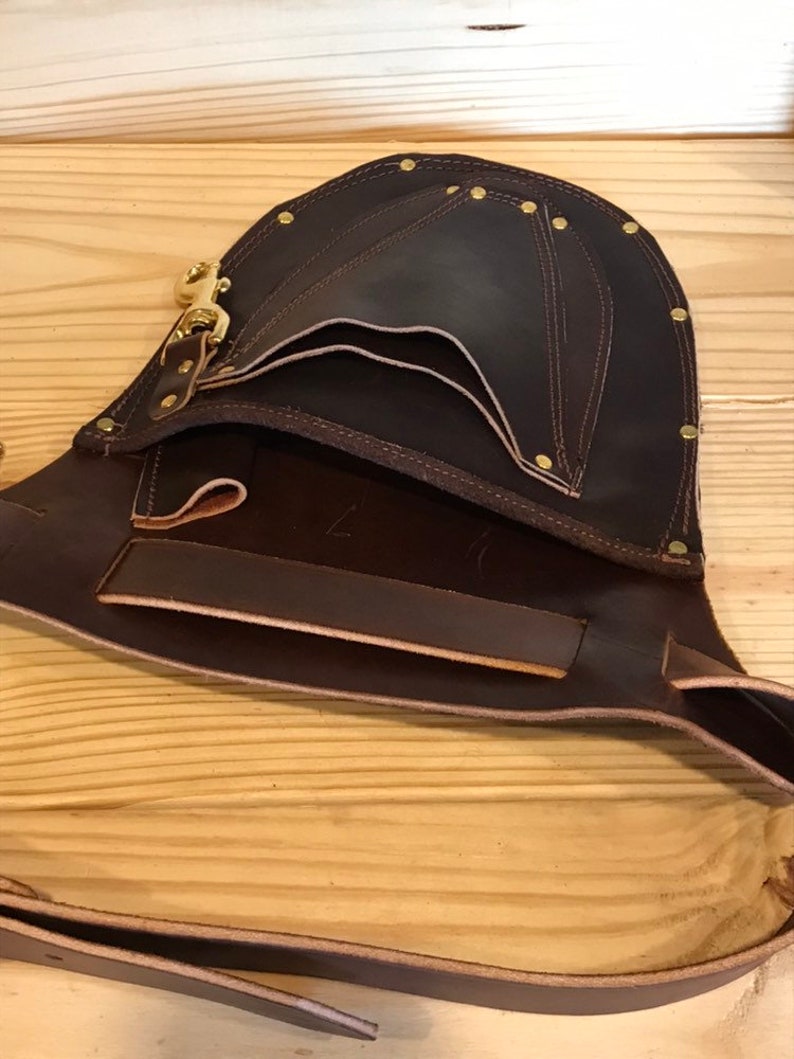 May include: A brown leather tool belt with multiple pockets and a brass buckle. The belt is open and the inside is visible.