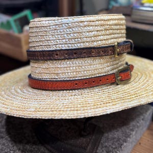 Puede incluir: Un sombrero de paja con ala ancha, con dos bandas de cuero en marr&oacute;n y naranja, cada una con una hebilla. El sombrero tiene una textura tejida y est&aacute; dise&ntilde;ado para la protecci&oacute;n solar. Las bandas de cuero a&ntilde;aden un elemento decorativo.