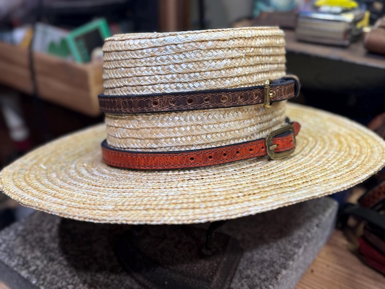 Puede incluir: Un sombrero de paja con ala ancha, con dos bandas de cuero en marr&oacute;n y naranja, cada una con una hebilla. El sombrero tiene una textura tejida y est&aacute; dise&ntilde;ado para la protecci&oacute;n solar. Las bandas de cuero a&ntilde;aden un elemento decorativo.