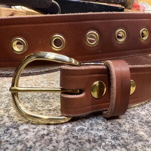 May include: A brown leather belt with a brass buckle and brass eyelets. The belt is thick and appears to be handmade. The buckle is a classic design, and the belt has a rugged, durable appearance.