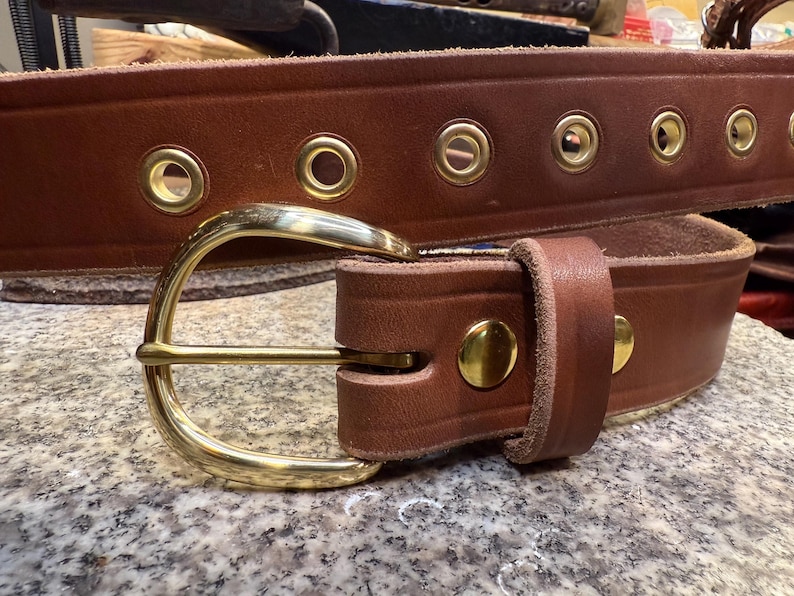 May include: A brown leather belt with a brass buckle and brass eyelets. The belt is thick and appears to be handmade. The buckle is a classic design, and the belt has a rugged, durable appearance.