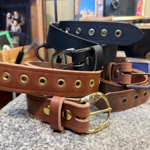 May include: Several leather belts in shades of brown and black are displayed. Some belts have metal eyelets and buckles in silver and gold. The belts are stacked on a textured surface, highlighting the craftsmanship.