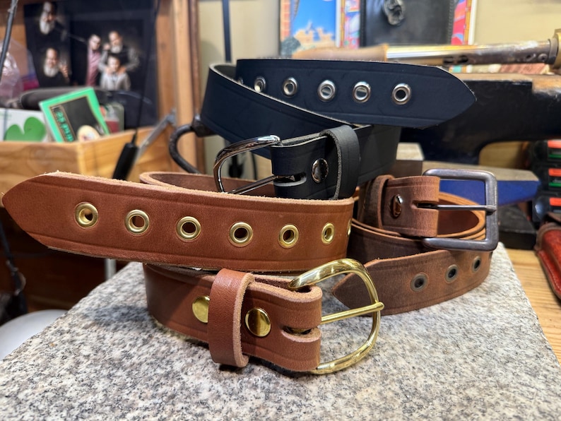 May include: Several leather belts in shades of brown and black are displayed. Some belts have metal eyelets and buckles in silver and gold. The belts are stacked on a textured surface, highlighting the craftsmanship.