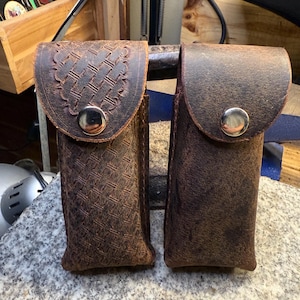 May include: Two brown leather pouches with snap closures. One pouch features a detailed woven pattern on the flap, while the other has a smooth, textured surface. Both are designed to hold small items.
