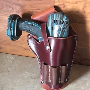 May include: A brown leather tool belt with a pocket for a drill and a smaller pocket with three slots. The drill is blue and black and is partially inserted into the belt.