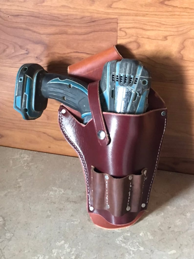 May include: A brown leather tool belt with a pocket for a drill and a smaller pocket with three slots. The drill is blue and black and is partially inserted into the belt.