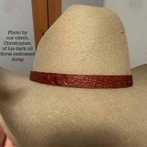 May include: A tan cowboy hat with a dark oil floral embossed strap. The hat has a classic western design, with a wide brim and a high crown. The strap is a rich, dark red color.