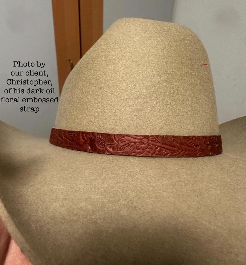 May include: A tan cowboy hat with a dark oil floral embossed strap. The hat has a classic western design, with a wide brim and a high crown. The strap is a rich, dark red color.