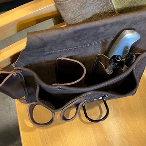 May include: A brown leather tool belt with multiple pockets and loops. The belt has a silver utility knife and two pens in the top pocket.