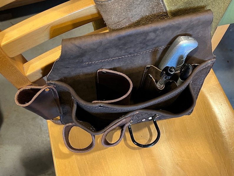 May include: A brown leather tool belt with multiple pockets and loops. The belt has a silver utility knife and two pens in the top pocket.