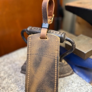 Op de afbeelding: Een bruine leren bagagelabel met een gestikte rand en een bijpassende leren riem. Het label heeft een verweerde uitstraling en een metalen gesp. De achtergrond is wazig, wat duidt op een werkplaats.
