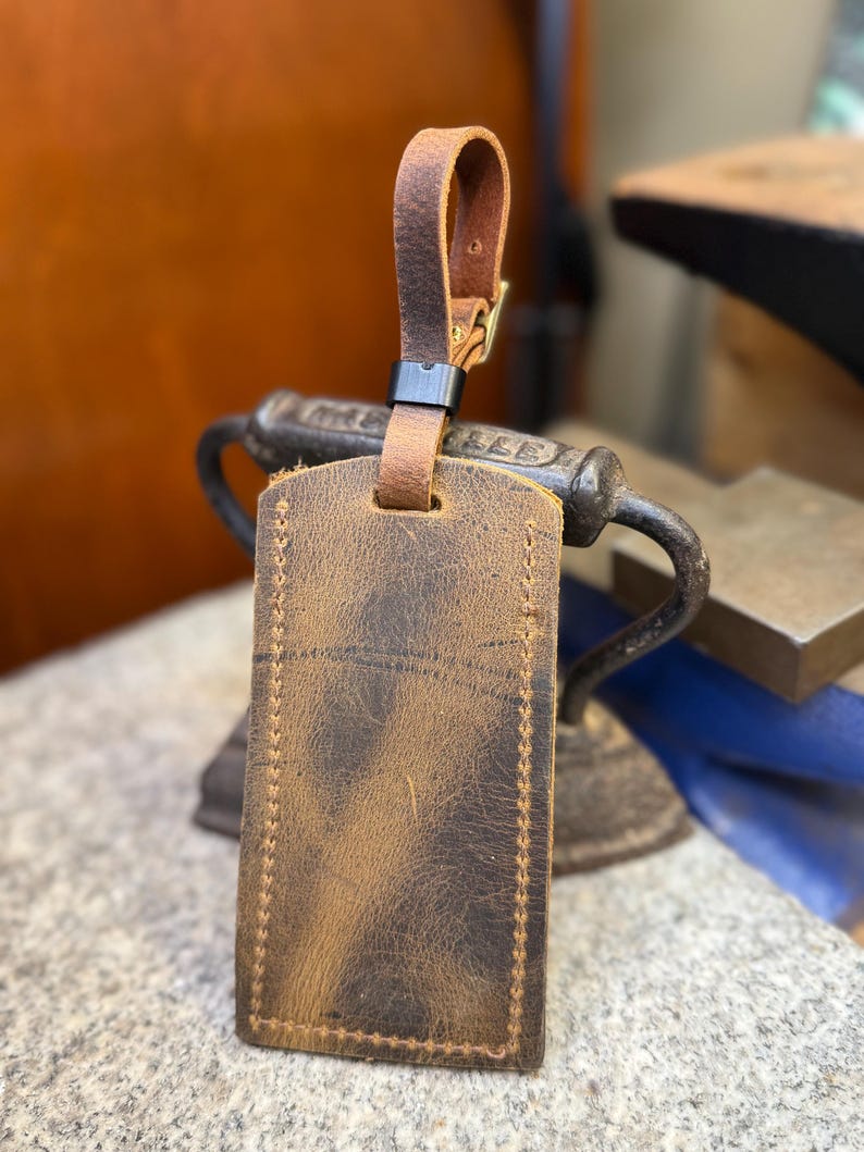 Op de afbeelding: Een bruine leren bagagelabel met een gestikte rand en een bijpassende leren riem. Het label heeft een verweerde uitstraling en een metalen gesp. De achtergrond is wazig, wat duidt op een werkplaats.