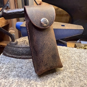 K&ouml;nnte beinhalten: Eine braune Lederscheide mit einer Klappe, die mit einem silbernen Knopf befestigt ist. Die rechteckige Scheide scheint handgefertigt zu sein. Das Leder hat einen Used-Look. Im Hintergrund sind ein B&uuml;geleisen und ein Amboss zu sehen.