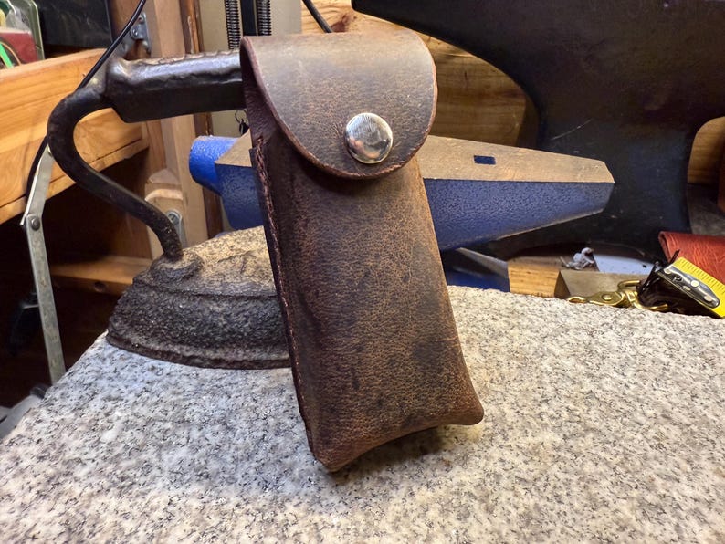 K&ouml;nnte beinhalten: Eine braune Lederscheide mit einer Klappe, die mit einem silbernen Knopf befestigt ist. Die rechteckige Scheide scheint handgefertigt zu sein. Das Leder hat einen Used-Look. Im Hintergrund sind ein B&uuml;geleisen und ein Amboss zu sehen.