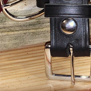 Peut inclure: Une ceinture en cuir noir avec une boucle argent&eacute;e et un bouton argent&eacute;. La ceinture est sur une surface en bois.